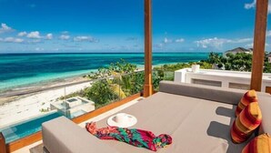 Un balcón con vista al océano y la playa, un rincón de descanso con una mesa y cojines, y un cielo despejado.