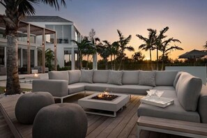 Uma área de estar moderna ao ar livre, com lareira, piscina e palmeiras ao fundo.