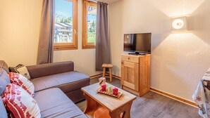 A cozy living room with a sofa, a small table with a tray of snacks, a television on a wooden cabinet, and two windows with curtains.