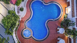 An aerial view of a swimming pool with a unique wavy edge design, surrounded by a red-tiled deck and outdoor seating with umbrellas.