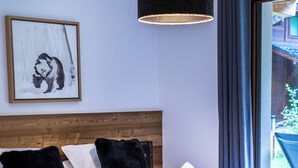 Een slaapkamer met een groot bed, een houten hoofdeinde, een zwarte hanglamp, een ingelijste foto aan de muur en uitzicht op het omliggende landschap door een raam.