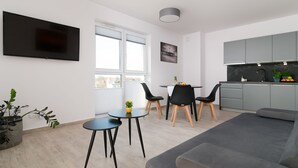 Una sala de estar moderna con un sofá gris, un televisor de pantalla plana montado en la pared, un comedor con una mesa pequeña y sillas, y una cocina con armarios grises y una encimera negra.