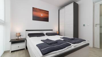 Quarto moderno com uma cama grande, mesa de cabeceira com abajur, guarda-roupa e uma pintura com pôr do sol na parede.