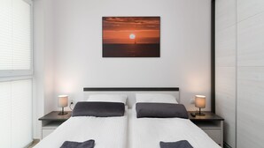 Habitación de hotel con dos camas, una mesita de noche con lámpara, un cuadro colgado en la pared de un atardecer y una ventana con persianas.