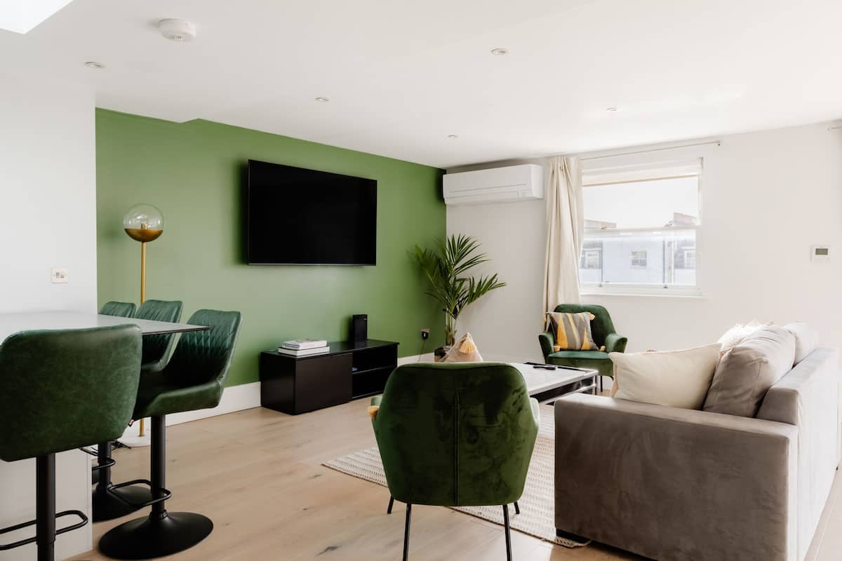 A modern living room with a green accent wall, a flat-screen TV, a green chair, a grey sofa, and a dining area with a table and chairs.