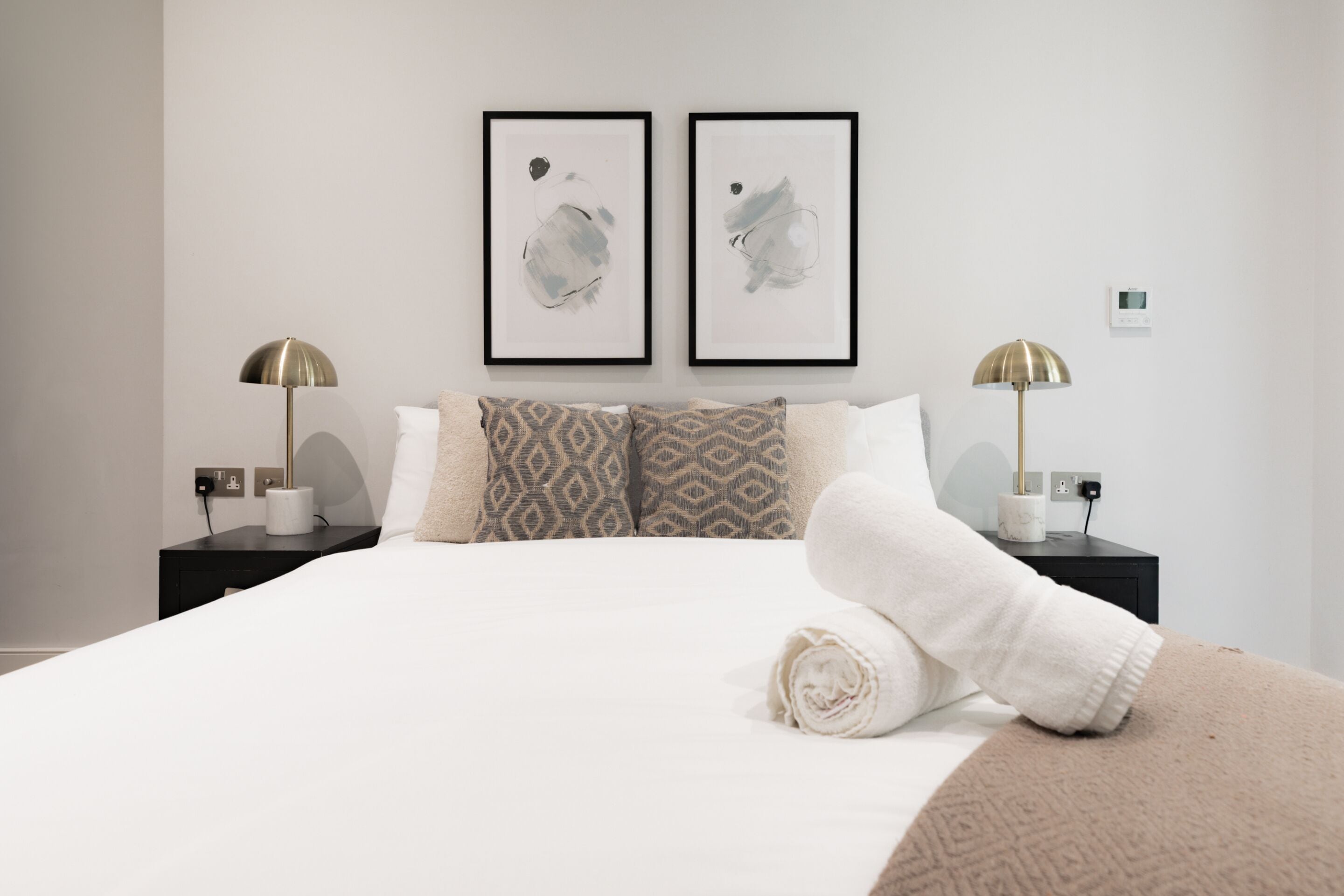 A neatly made bed with a rolled towel, two framed abstract artworks on the wall, and two bedside lamps on black nightstands.