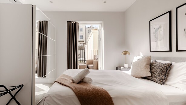 Une chambre d’hôtel moderne, dotée d’un grand lit, offrant une vue sur la ville grâce à une porte ouverte, et présentant une décoration minimaliste.
