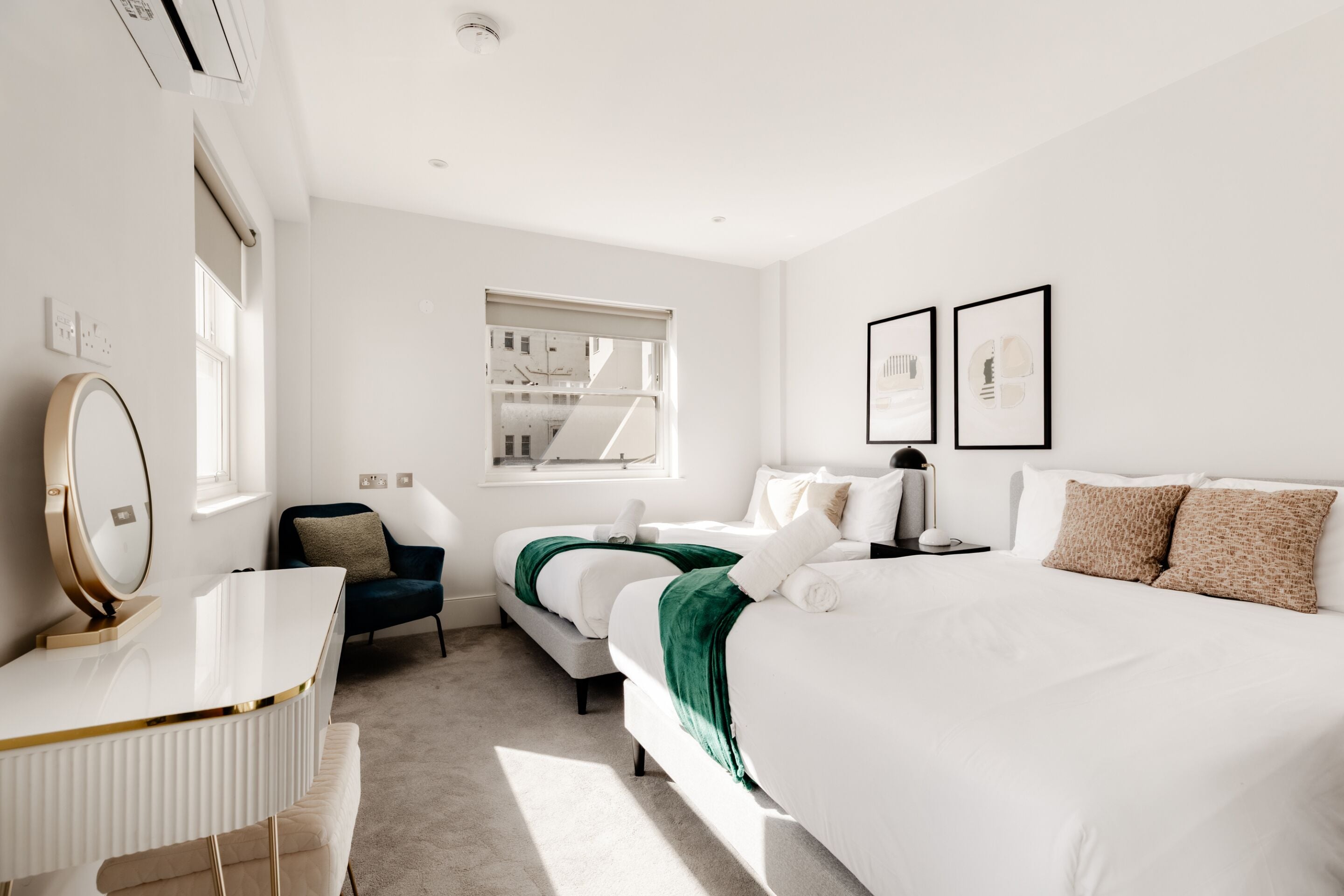 Ein modernes Hotelzimmer mit einem großen Bett, einem Schreibtisch mit Spiegel, einem Sessel und einem Fenster mit Blick auf das Gebäude.