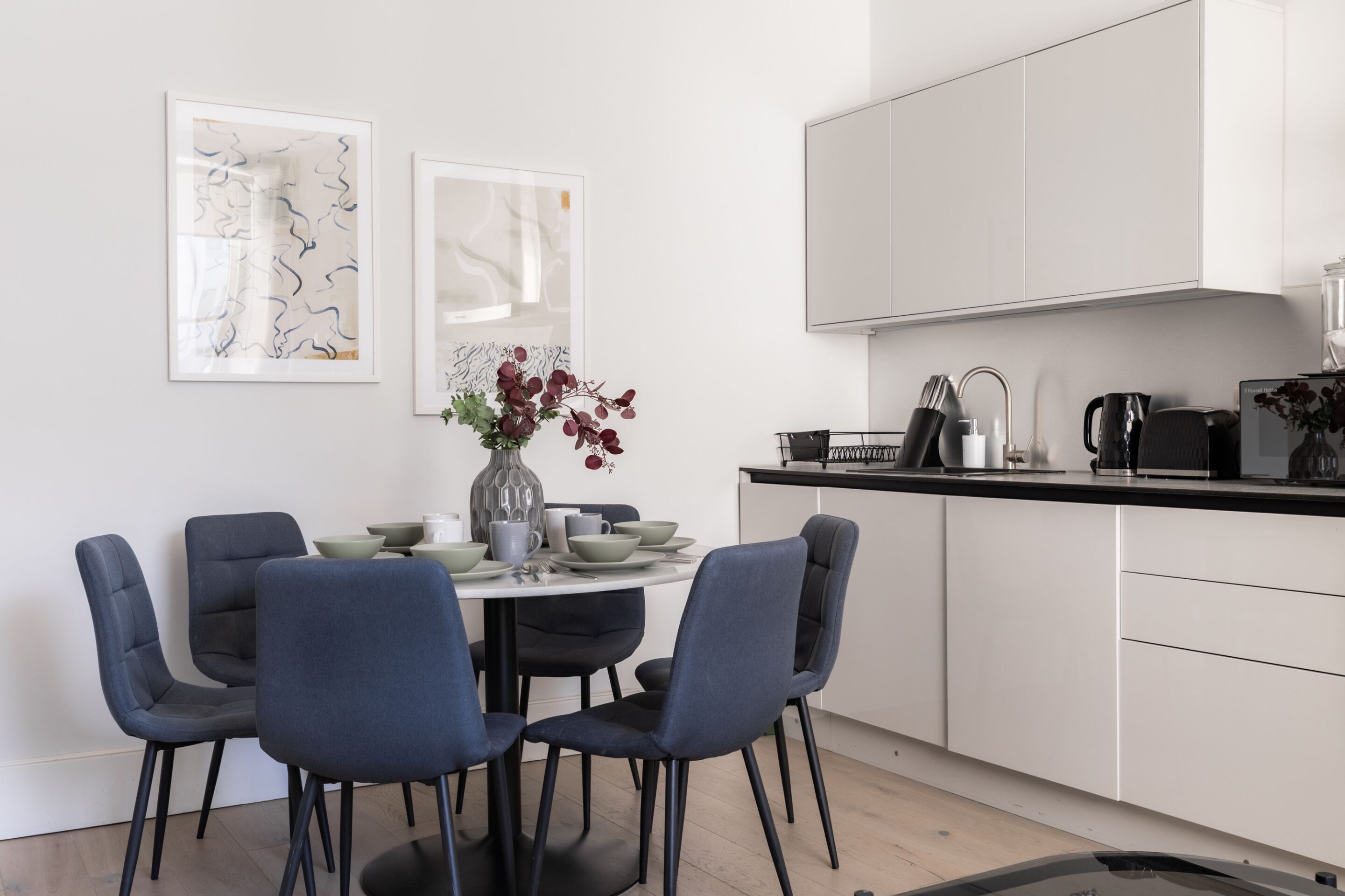 A modern kitchen with white cabinets, a black countertop, and a dining area with blue chairs and a white table.