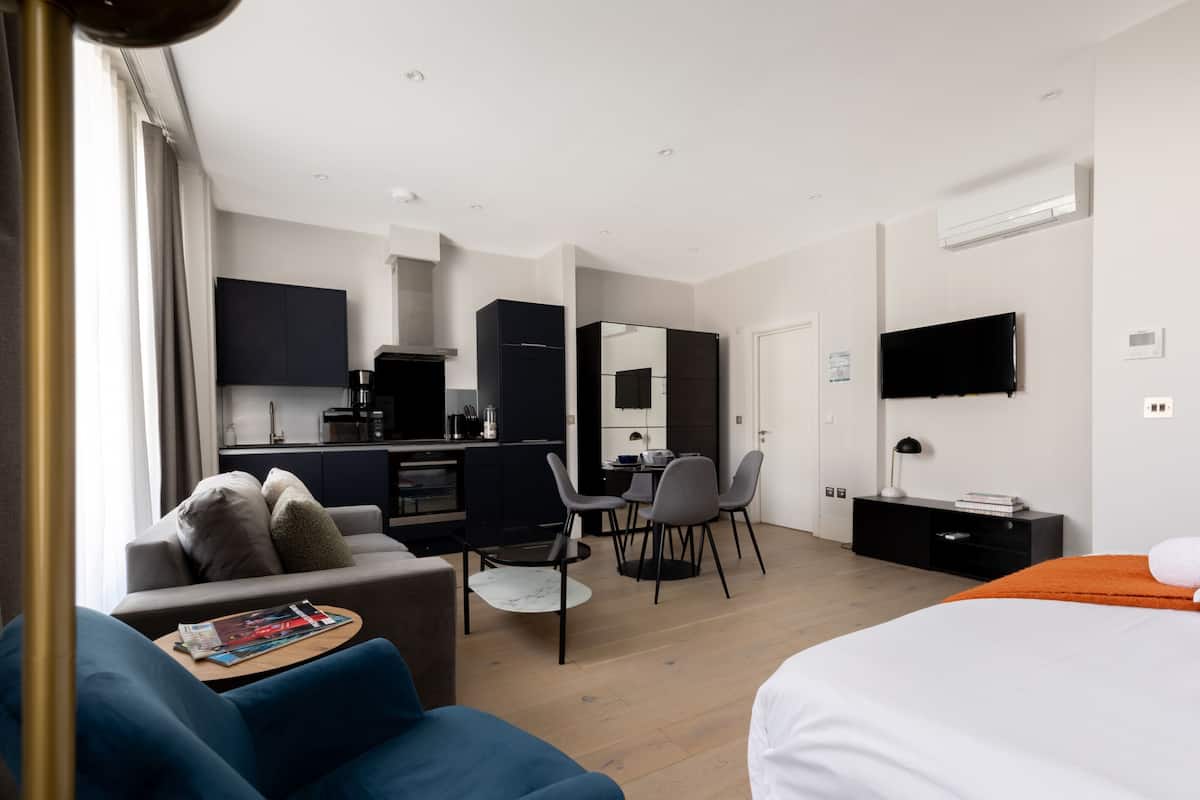 A modern hotel room with a sofa, a bed, a dining area with chairs, a kitchen area, and a TV mounted on the wall.