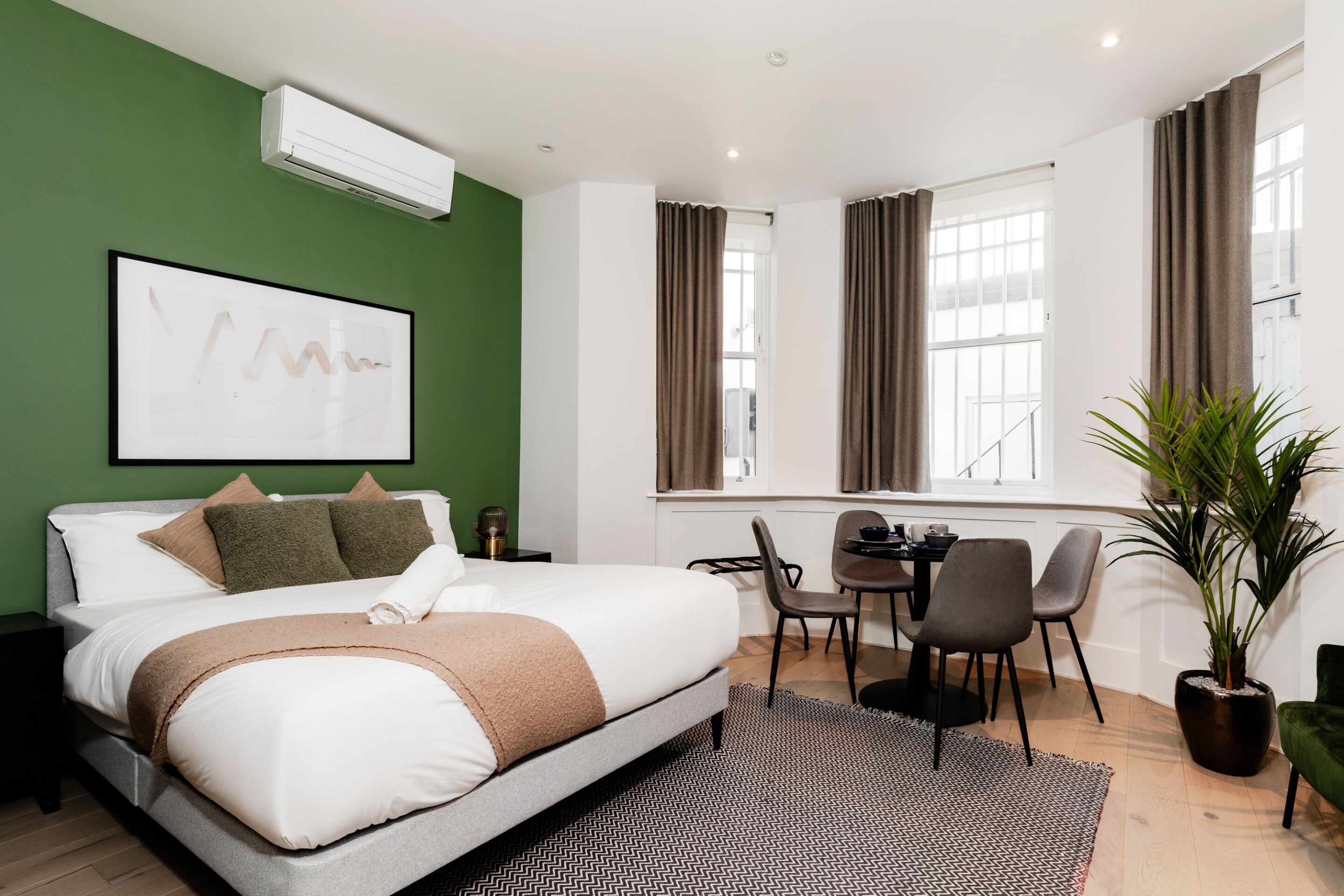 A modern hotel room with a large bed, a dining area, and a potted plant.