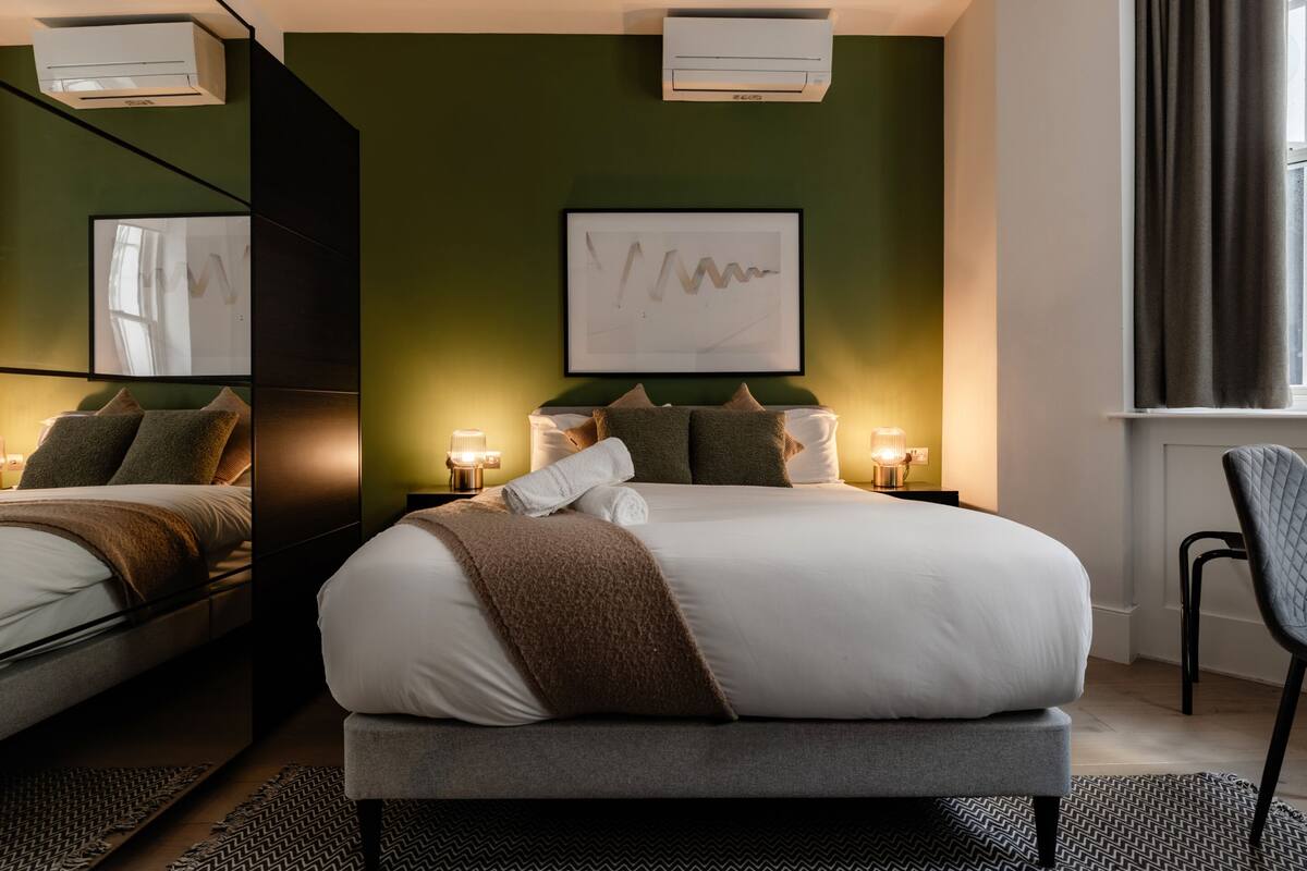 A modern hotel room with a large bed, bedside tables with lamps, a framed artwork on the wall, and an air conditioning unit.