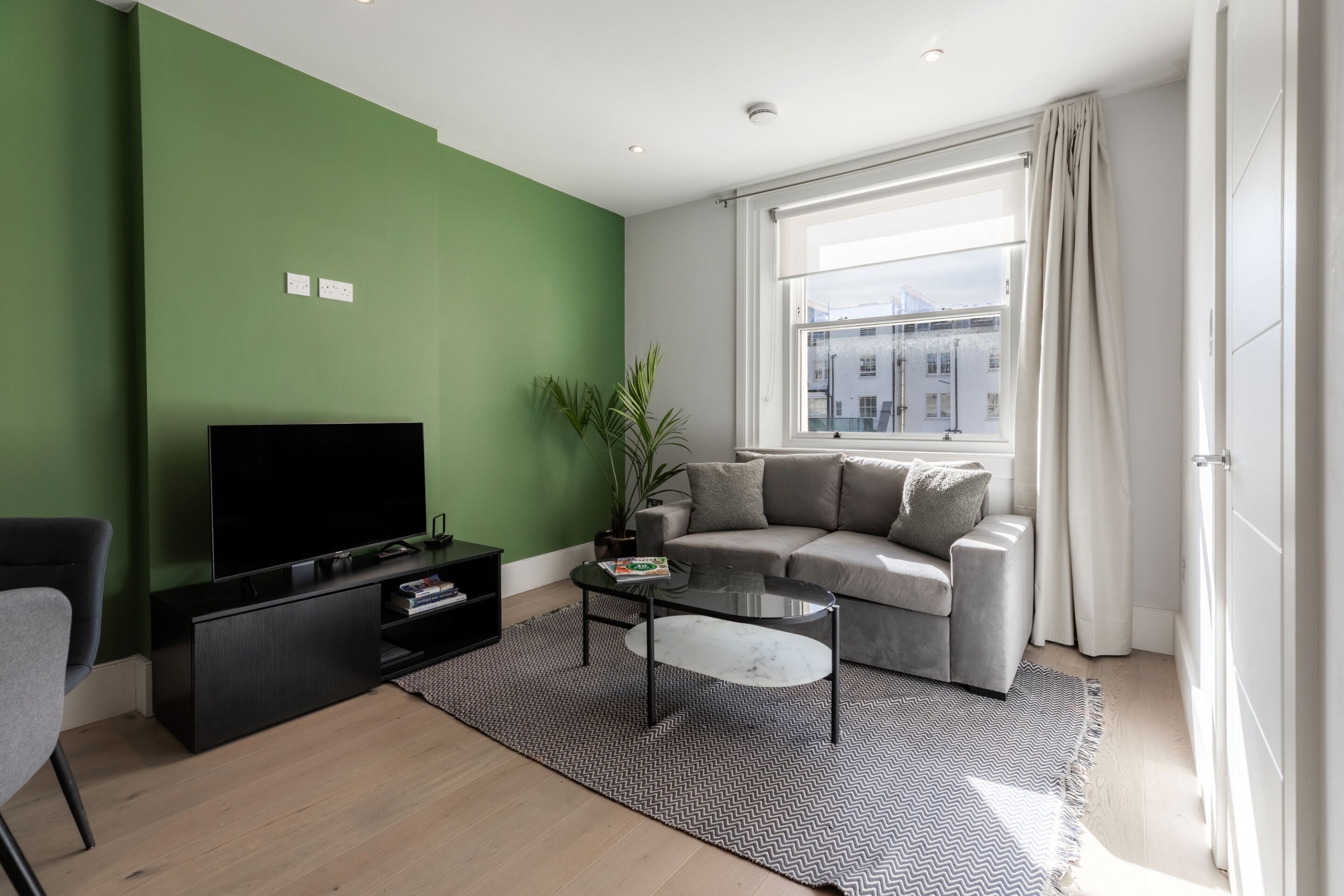 A modern living room with a green wall, a grey sofa, a black entertainment unit, and a large window with curtains.