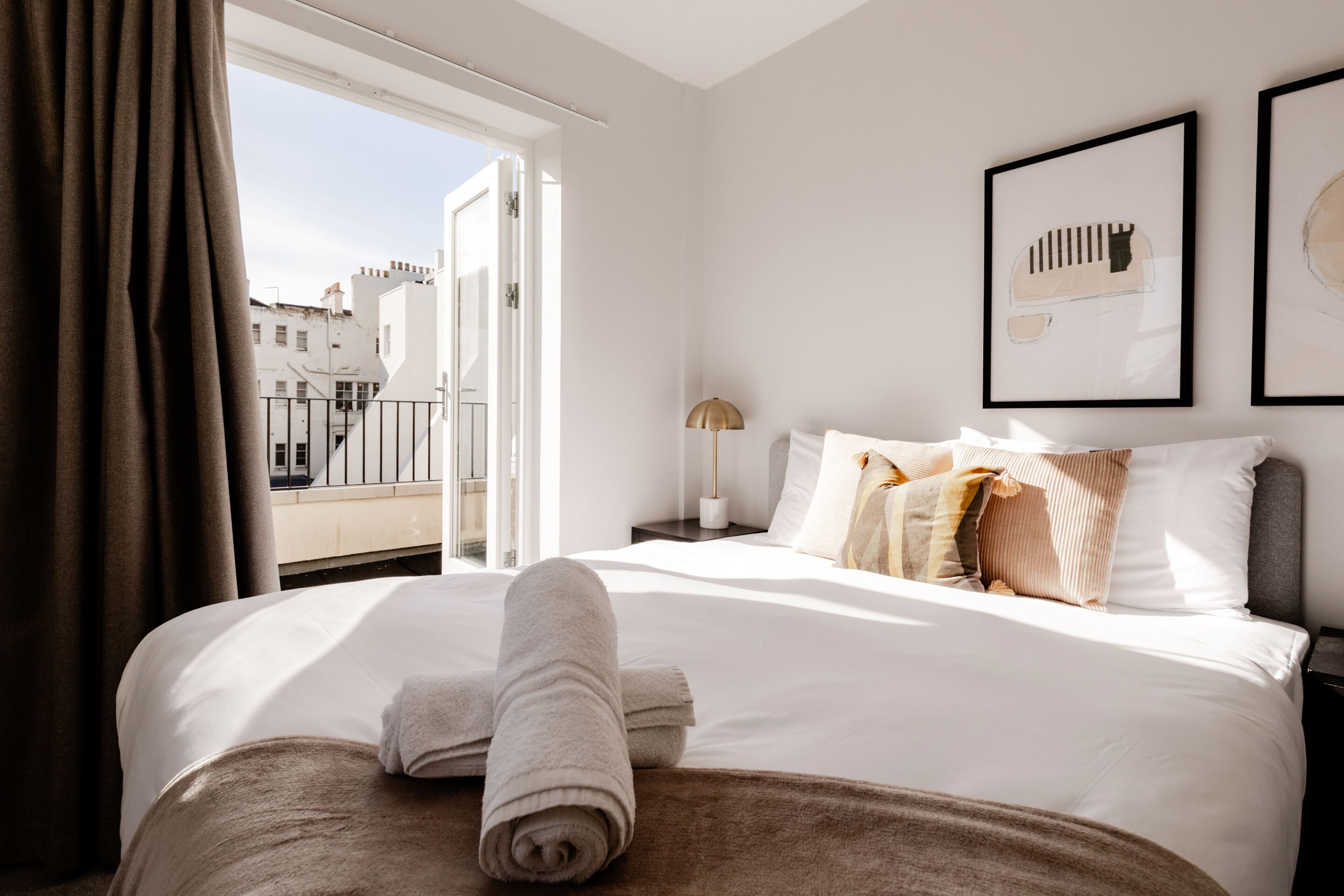 Une chambre d’hôtel avec un lit, une fenêtre avec des rideaux, une table de chevet et des œuvres d’art encadrées accrochées au mur.