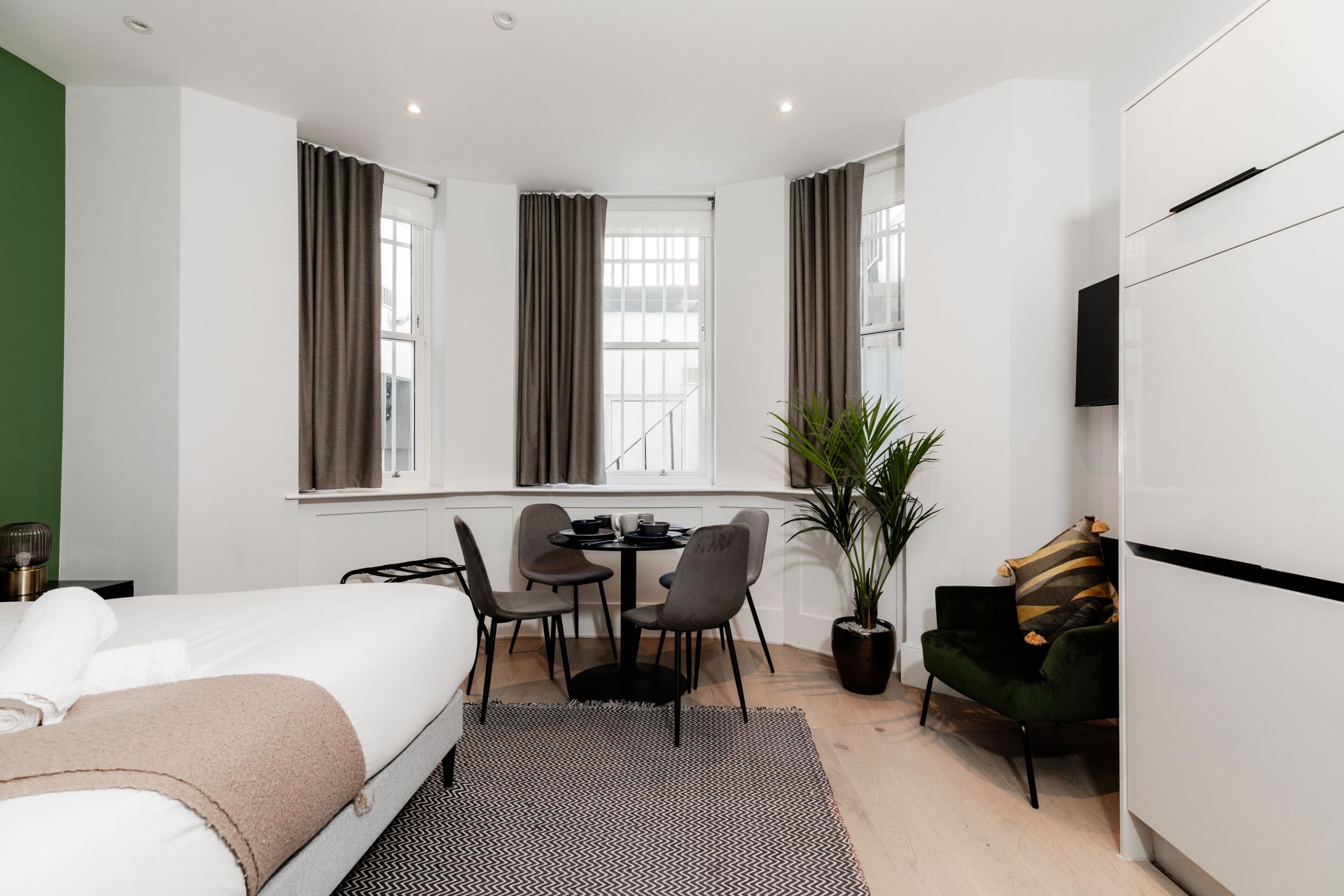 A modern hotel room with a bed, a desk with chairs, a potted plant, and a TV on the wall.