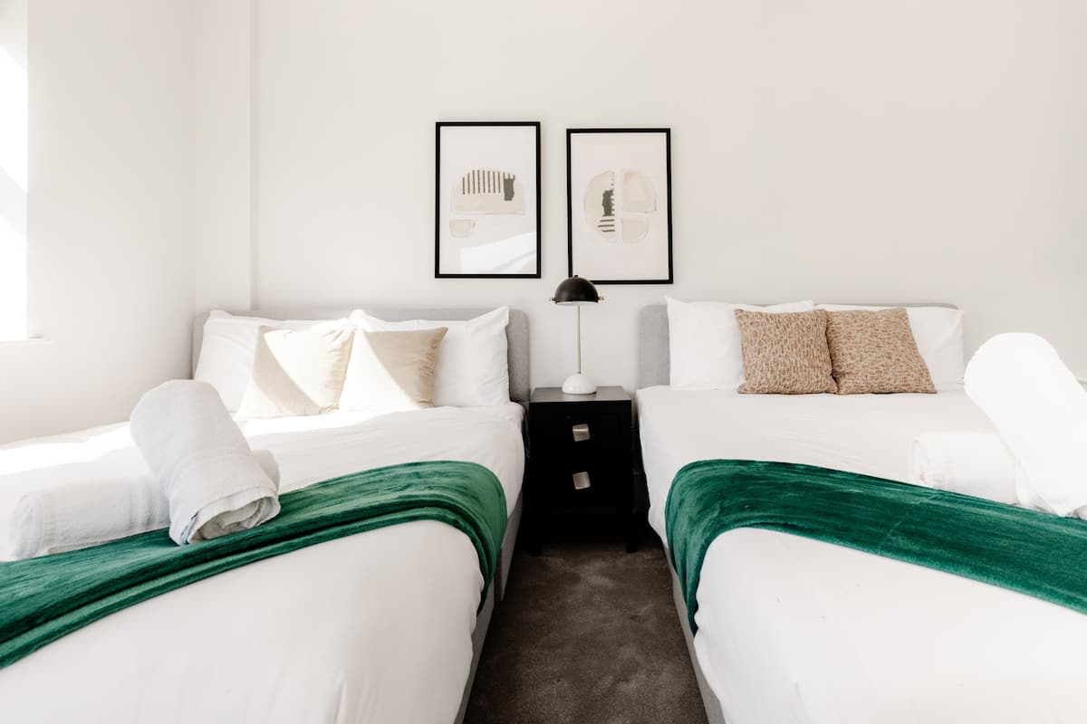 A hotel room with two beds, white bedding, green throw blankets, a nightstand with a lamp, and two framed artworks on the wall.