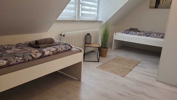 A compact, modern bedroom with a sloped ceiling, a single bed with a patterned bedspread, a small side table with a plant, and a white sofa. - Apartment COLIN (Bremerhaven)