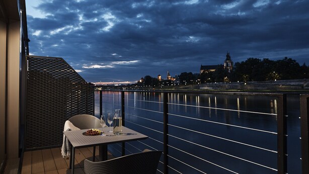 A balcony with a table set for two, overlooking a river with a cityscape in the background. - New Port Hotel (Kraków)