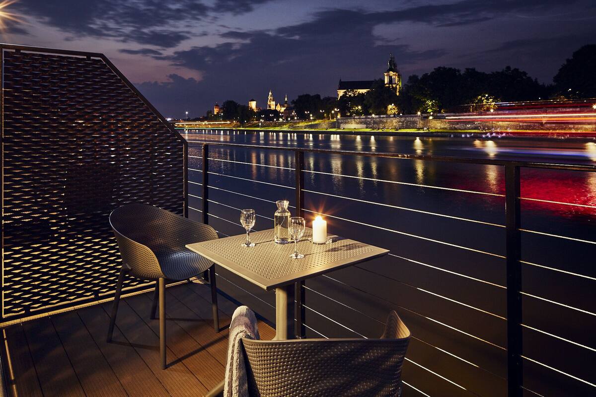 An outdoor dining area with a table and chairs, a candle, and a view of a river with city lights in the background.