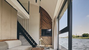 A modern interior with a sofa, two small tables, a wooden accent wall, a TV, and a view of a river. - New Port Hotel (Kraków)