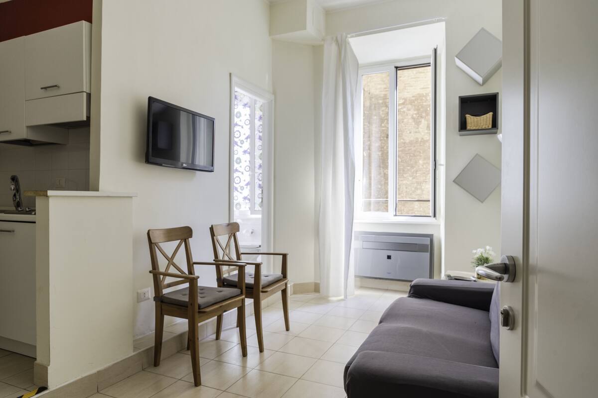 A modern living room with a sofa, two wooden chairs, a flat-screen TV, and a window with curtains.