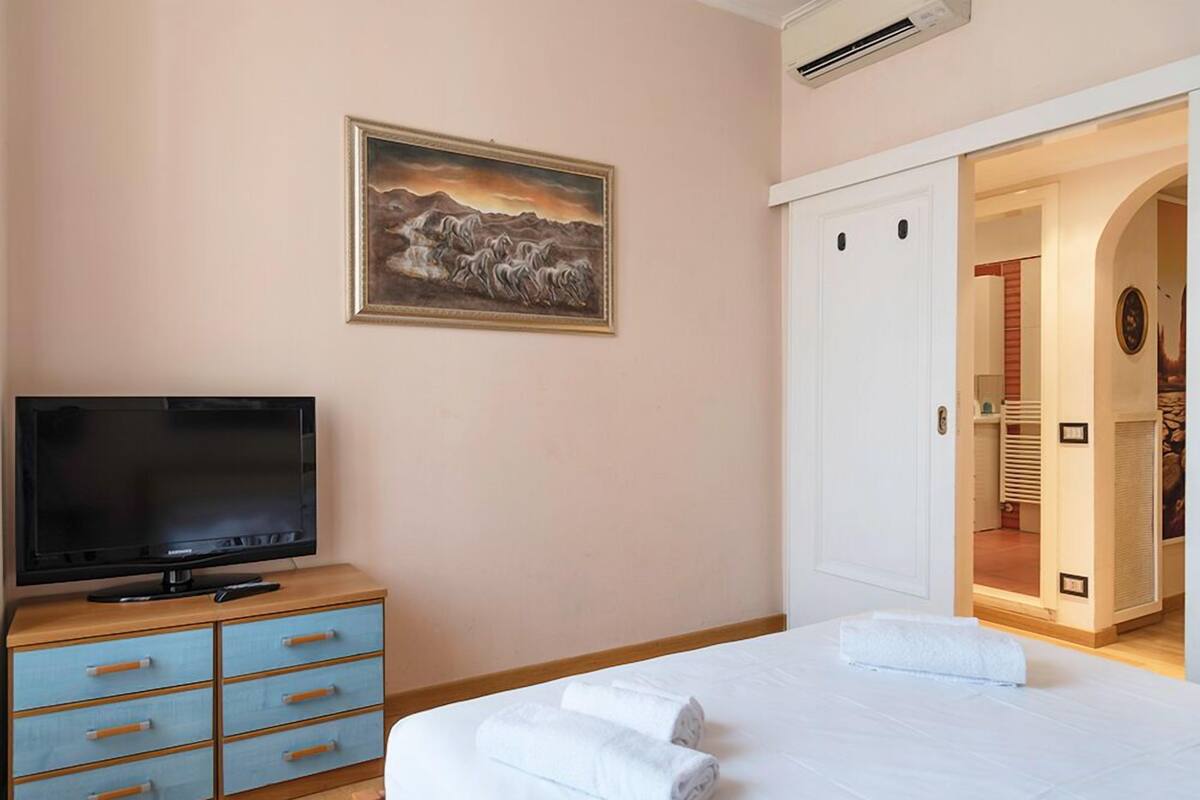 A bedroom with a bed, a TV on a wooden dresser, a painting on the wall, and a wardrobe.