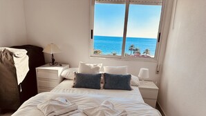 Une chambre à coucher avec un lit, des oreillers, une table de chevet, une lampe et une vue sur la mer par la fenêtre.