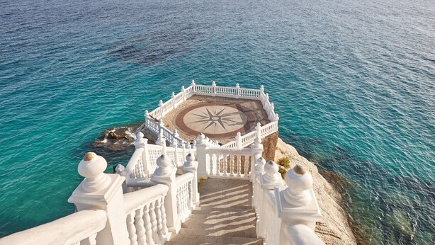 Un escalier blanc menant à une plateforme circulaire ornée d’un motif en forme de boussole, d’où l’on peut admirer une mer turquoise.