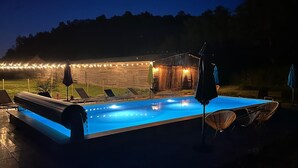 Piscina com espreguiçadeiras e um pequeno edifício ao fundo, decorado com luzes de corda.