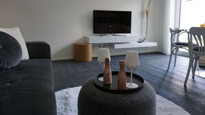 A modern living room with a dark grey sofa, a black ottoman with decorative bottles and glasses, a flat-screen TV on a white console, and a white chair by a window. - Ferienhaus mit 6 Apartments, Terrasse und Blick auf den Stausee (Bad Lobenstein)