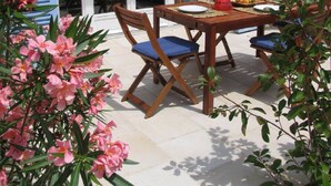 Une table en bois, dressée pour deux personnes, avec une chaise, une bouteille et des fruits, placée à l’extérieur d’un bâtiment aux volets bleus et aux murs de pierre.