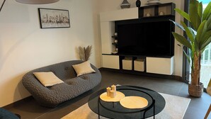 Una sala de estar moderna con un sofá gris, una mesa de centro negra, un televisor de pantalla plana y una planta grande en maceta.