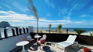 Una terraza con mesa y sillas, con vista a una playa con palmeras y un cielo despejado.