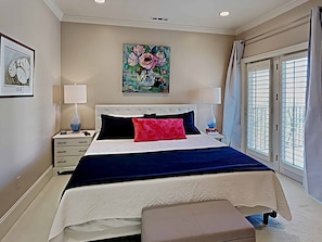 A bedroom with a large bed, white nightstands, a painting on the wall, and a window with curtains.