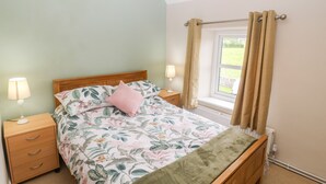 Une chambre à coucher avec une couvre-lit à motifs floraux, une tête de lit en bois, une table de chevet avec une lampe et une fenêtre avec des rideaux.