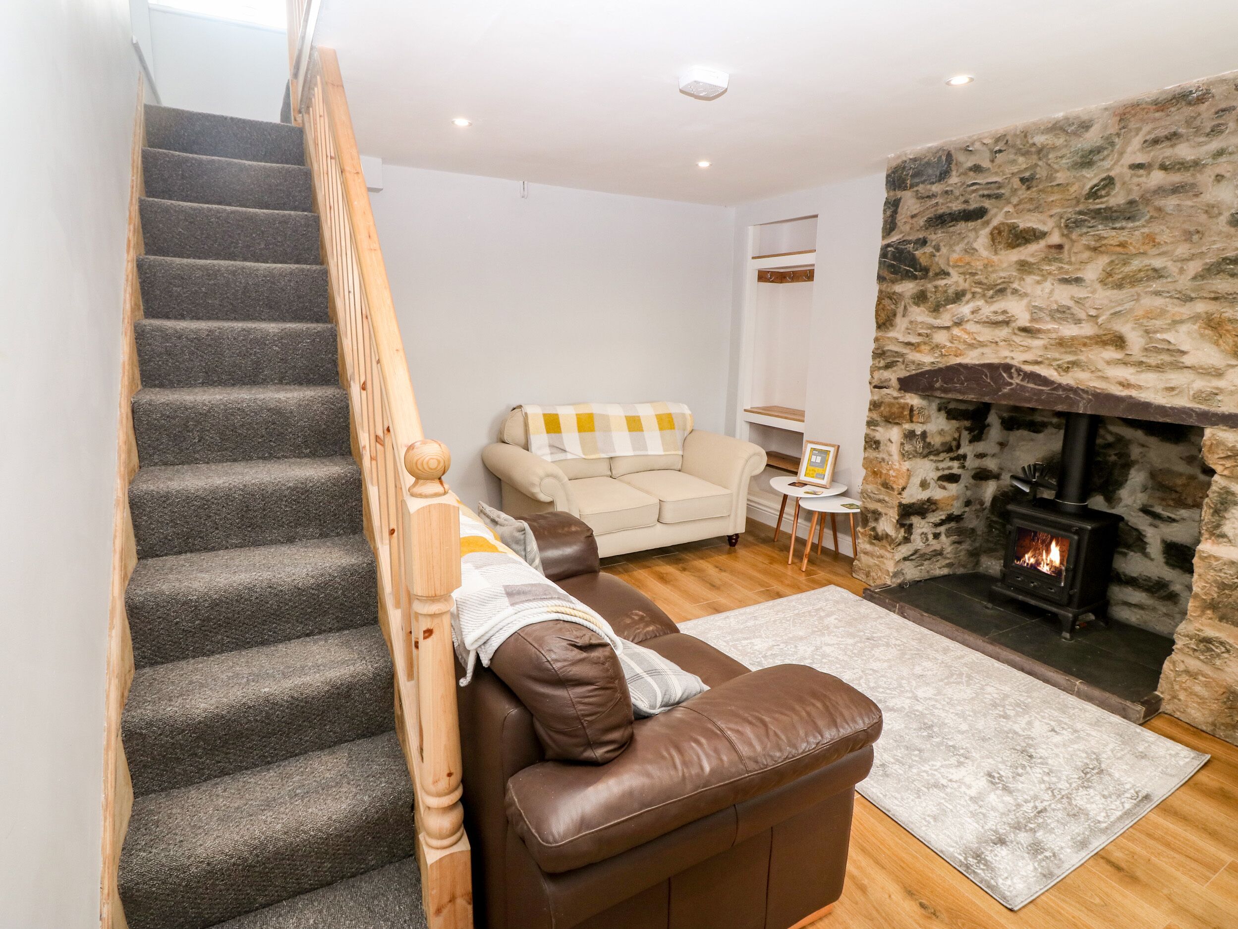 A staircase with wooden balusters, a beige sofa with a checkered blanket, a brown leather chair, a stone wall, and a wood-burning stove.