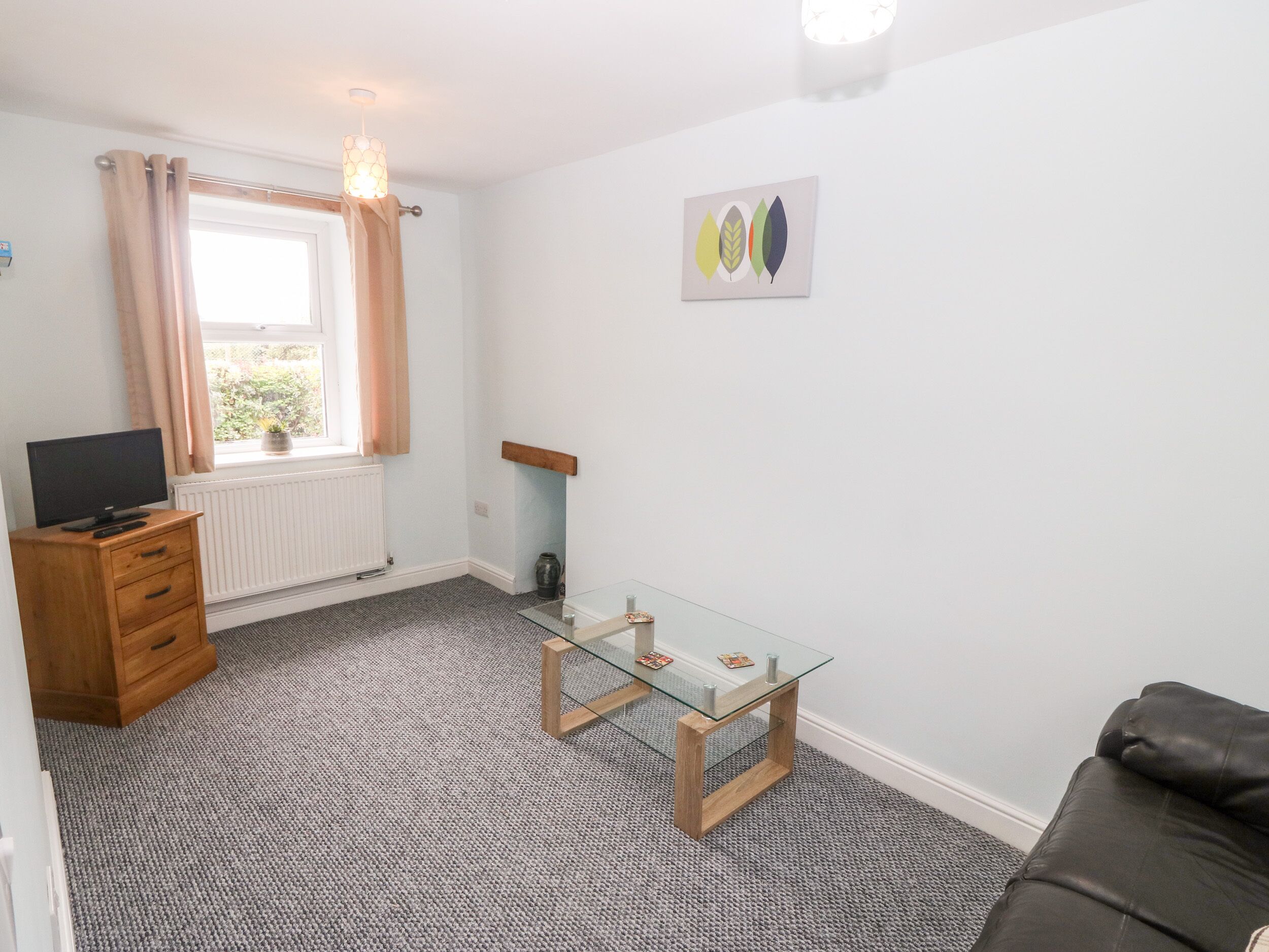 A small, tidy room with a wooden dresser, a glass table, a black leather sofa, a window with curtains, and a wall-mounted artwork.