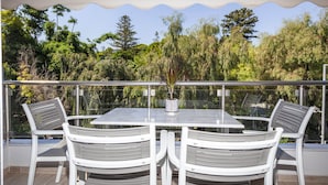 Un espace extérieur abrité, aménagé pour les repas, avec une table et quatre chaises, entouré d’une rambarde en métal et donnant sur une zone boisée.