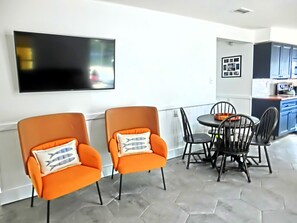 A modern dining area with two orange armchairs, a round table, and black chairs. A flat-screen TV is mounted on the wall above the seating area.