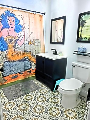 A bathroom with a shower curtain featuring a mermaid design, a white toilet, a sink with a black faucet, and a mirror above the sink.