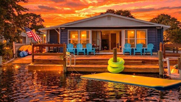 A house with a dock extending into a body of water, featuring a flag and outdoor seating. - Family & Pet-Friendly River Escape w/ Kayaks, Manatees & Outdoor Fun! (Weeki Wachee)