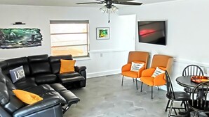 A living room with a black leather sofa, orange armchairs, a dining table with chairs, a ceiling fan, and artwork on the walls. - Family & Pet-Friendly River Escape w/ Kayaks, Manatees & Outdoor Fun! (Weeki Wachee)