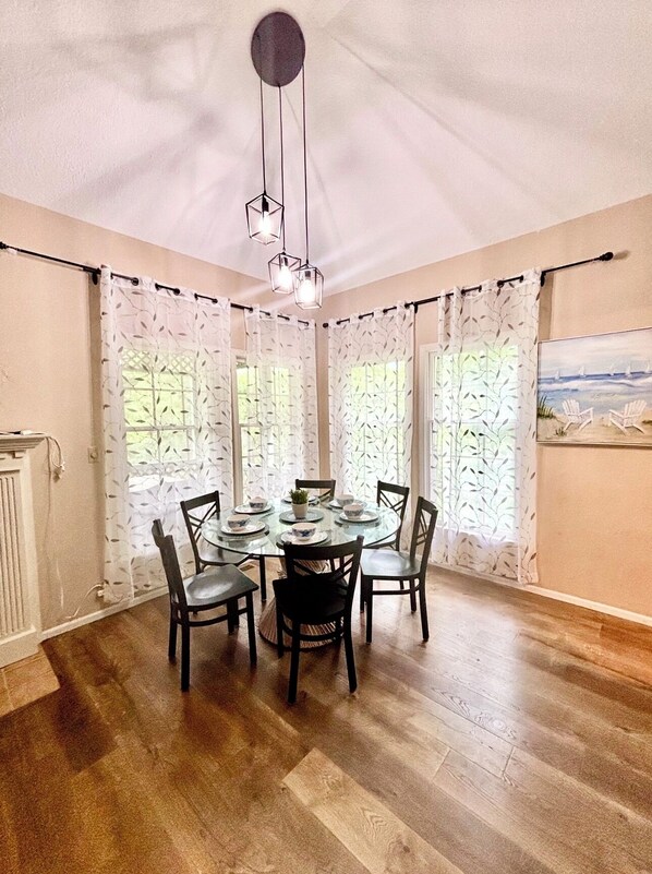 Un comedor con una mesa redonda, seis sillas, una lámpara de araña y cortinas estampadas.