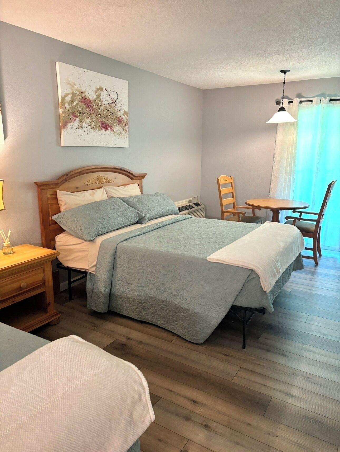 A bedroom with a bed, bedside table, chair, and a painting on the wall.