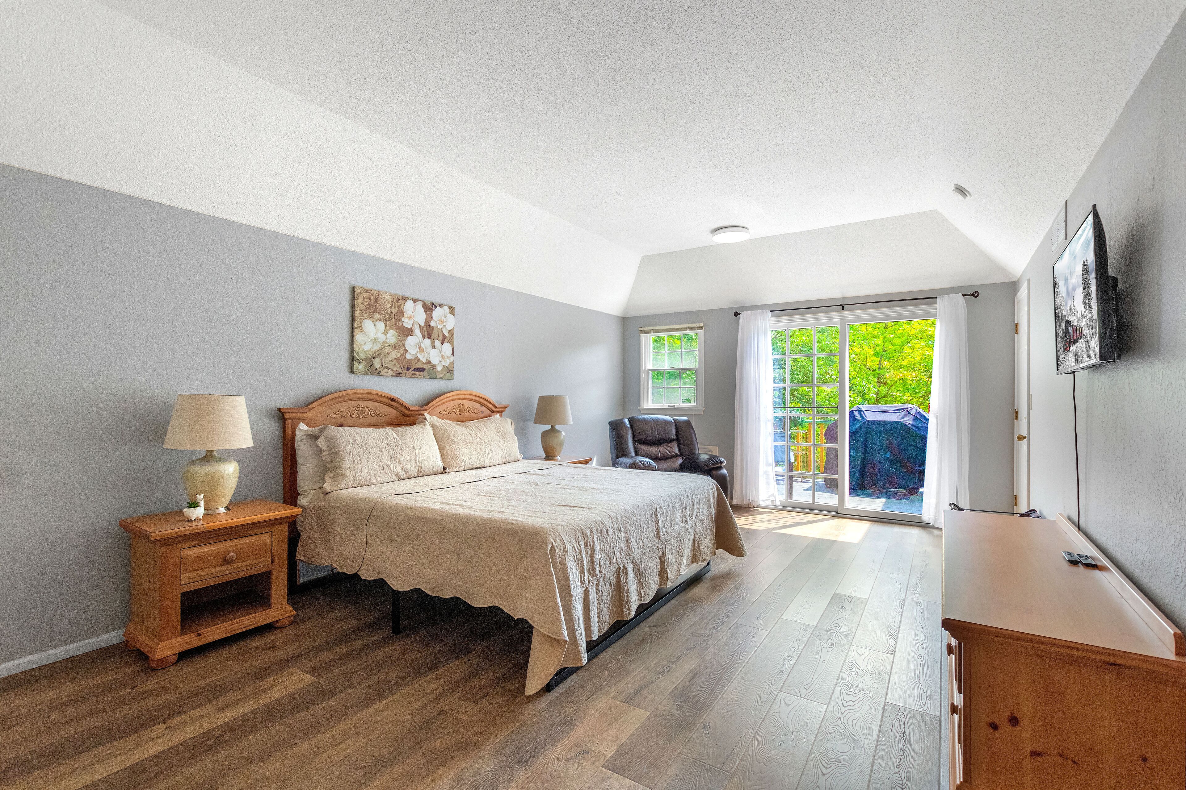A bedroom with a bed, nightstands, a chair, a TV, and a view of greenery outside.