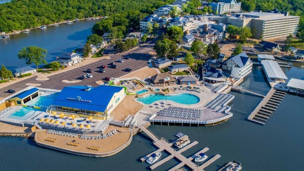 An aerial view of a lakeside resort with a large swimming pool, multiple docks, and a variety of buildings including a hotel and a large resort building. - "The Mint" @ Margaritaville Lake Resort, Boating, Golfing, Fire Pit, Swimming (Osage Beach)