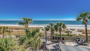 Blick auf einen Strand von einer Holzterrasse aus mit Liegestühlen und Palmen.