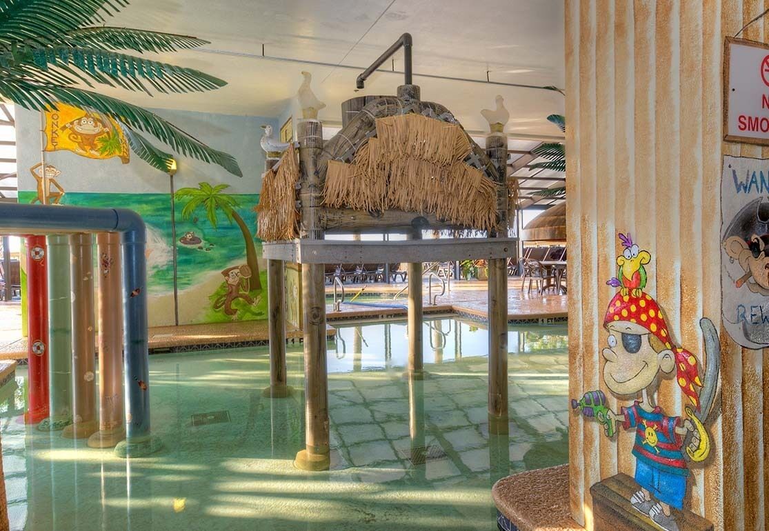 Un área de juegos acuáticos con un mural de una escena de playa, una estructura con techo de paja y un personaje de caricatura en la pared.