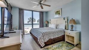 Ein Schlafzimmer mit Bett, Nachttischen, Deckenventilator, Fenster mit Vorhängen und Blick auf den Strand.