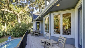 A covered outdoor deck with a pool, wooden furniture, and large windows overlooking a lush garden. - 10 Heath - Breathtaking Fazio Golf Course Views (Hilton Head Island)